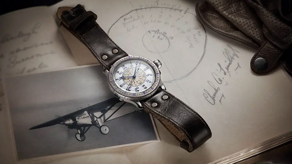 A vintage-style aviator watch with a silver case and cream dial displaying chronograph subdials, placed on a dark brown leather strap.