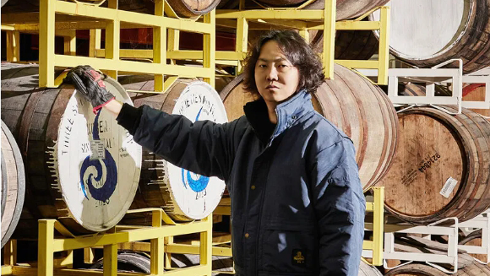 Mr. Kim Chang-soo wearing blue overalls standing in front a stack of whiskey barrels. The barrels are arranged on yellow metal racks in a well-lit warehouse.