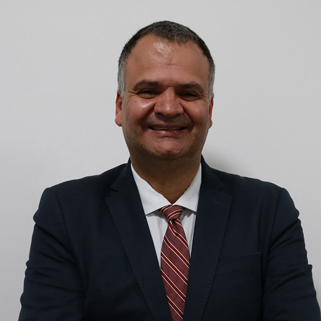 o Un homme vêtu d’un costume sombre, d’une chemise blanche et d’une cravate à rayures rouges, regarde directement l’appareil, les bras légèrement croisés. Son image se détache d’un fond blanc uni.