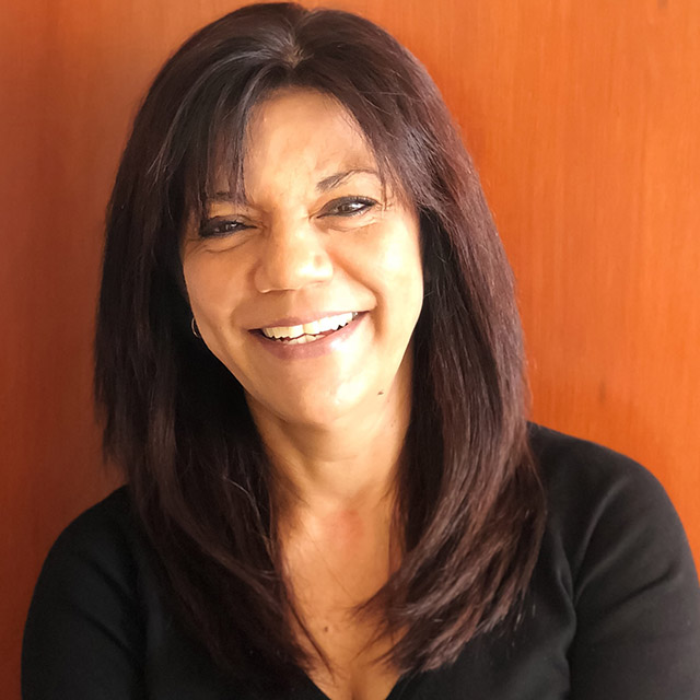 o Une femme aux cheveux bruns et raides, vêtue d’un haut noir, sourit à l’appareil, sur fond boisé.