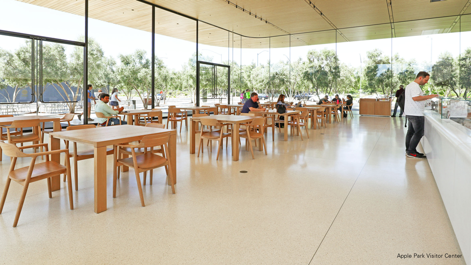 Photo shows the inside of a glass-fronted cafeteria with a low wooden ceiling. Nmerous HIROSHIMA chairs surround the cafeteria tables. A man in blue jeans and a white t-shirt is standing at a counter.