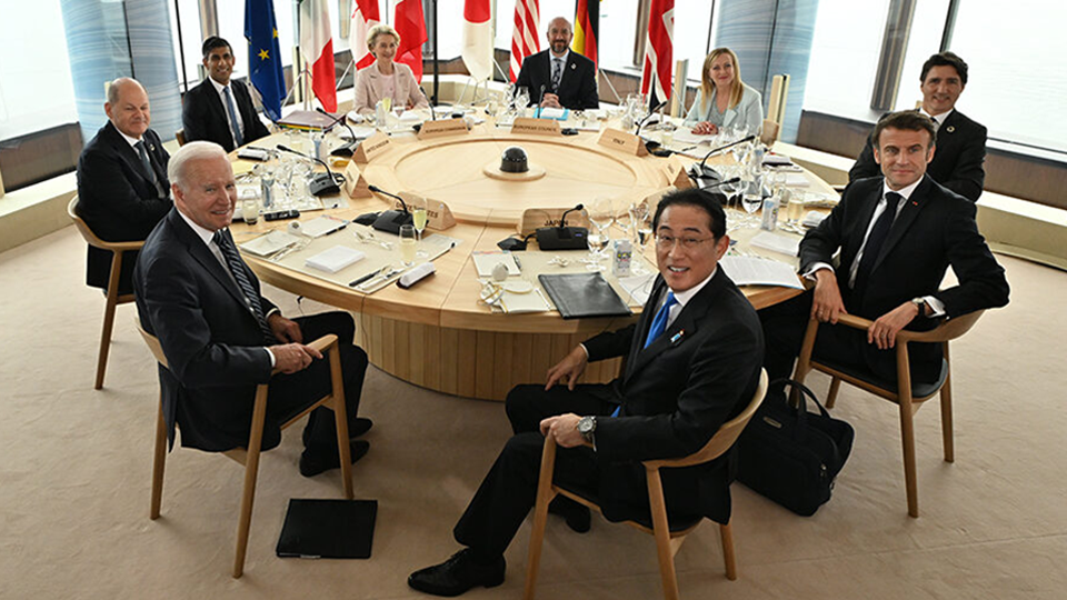 На фотографии показаны лидеры стран Группы семи, которые сидят на стульях HIROSHIMA за круглым деревянным столом, сзади них стоят национальные флаги.