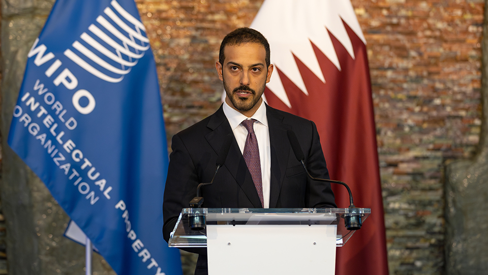 Ambassador Ahmed Essa Al-Sulaiti, Director of the Office of the State of Qatar to the World Trade Organization and Other Economic Organizations in Geneva, standing at a podium holding a speech. The WIPO and Qatari flags are behind him.