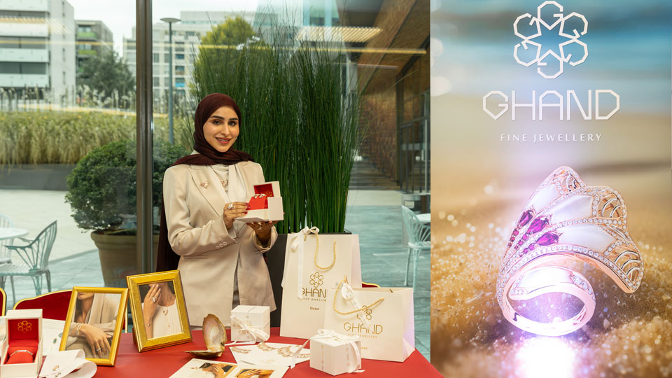 Photo shows Ms. Jawaher Mohammed Al Mannai, wearing a cream-colored abaya and deep red head scarf, standing behind a display table holding a box containing GHAND jewelry. The table is covered with gift bags, framed photos of products, and samples of jewelry. A display panel shows a gold and diamond encrusted ring and GHAND’s logo. Text reads GHAND Fine Jewellery.