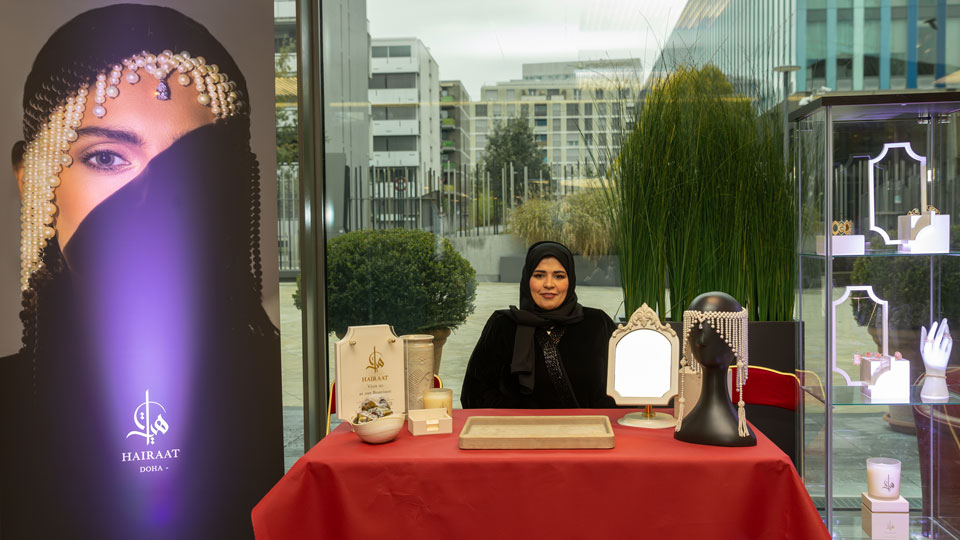 Photo of Ms. Nada Al Sulaiti wearing a black abaya, sitting behind a table covered with red drapes; a glass showcase containing samples of Hairaat’s products including rings, bracelets and necklaces. A publicity panel shows a model wearing a Hairaat pearl and diamond headdress.