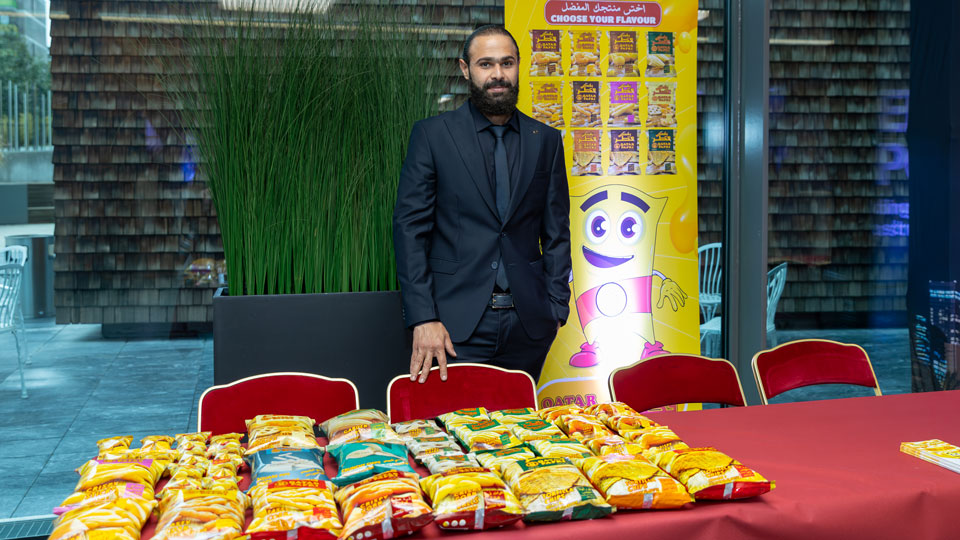 Photo shows Mr. Mazen Gabr Fayad wearing a black suit, shirt and tie, standing behind a display table and four red chairs. The table is covered with packets of QATAR PAFKI snacks in orange, green and red packets. A yellow display panel features photos of different packets of QATAR PAFKI snacks. Text reads Choose your flavour.