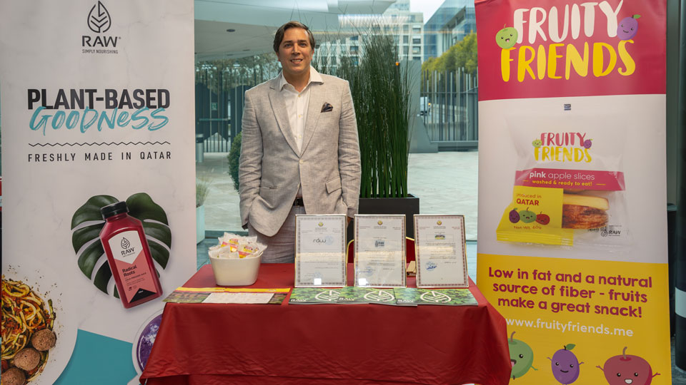 Photo shows Mr. Gabe Glennon wearing a grey suit and open neck white shirt, standing behind a display table with framed copies of RAW’s national trademark registrations and publicity materials. A white display panel features the RAW logo and a bottle of RAW Radical roots – a cold-pressed juice – lying on a green leaf. Text reads RAW Simply Nourishing, Plant-based goodness, Freshly made in Qatar. A second panel shows the Fruity Friends brand and a packet of ‘pink apple slices’. Text reads Low in fat and a natural source of fiber – fruits make a great snack.